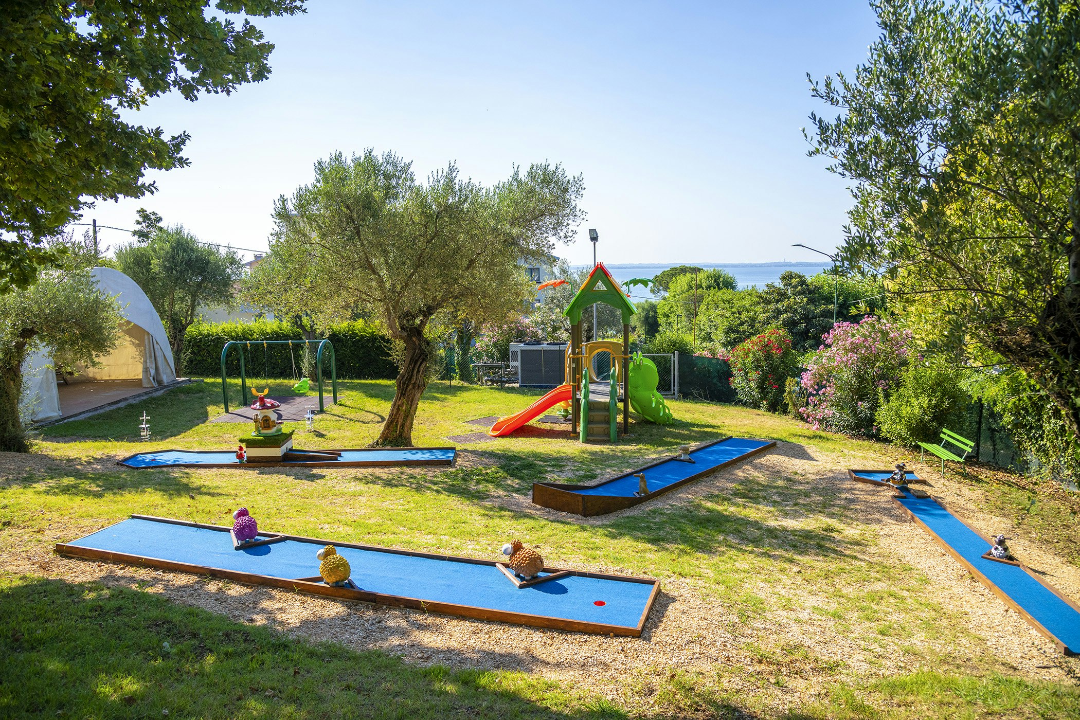 Spielbereich mit mehreren Minigolfbahnen und Trampolin im Campinggarten, umgeben von Rasen, Bäumen und Blick auf das Meer.