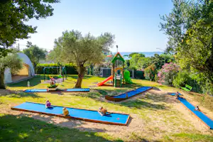 Spielbereich mit mehreren Minigolfbahnen und Trampolin im Campinggarten, umgeben von Rasen, Bäumen und Blick auf das Meer.