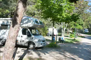 Wohnmobil auf dem Campingplatz, Schatten unter Bäumen, im Hintergrund weitere Fahrzeuge und Campingstühle am Weg.