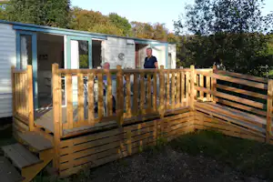 Mobilheim mit Holzterrasse, Blick auf die Eingangstür und Fenster, im Hintergrund Bäume und Hecken.