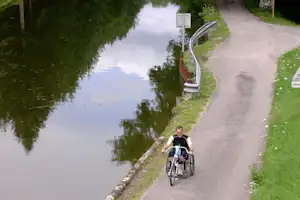 Radfahrer fährt auf dem Weg am Flussufer entlang, mit Geländer und Grasstreifen neben dem Wasser.
