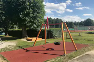 Spielplatz mit roter Holzschaukel auf rotem Boden, Hecken, Bäumen und eingezäuntem Sportfeld im Hintergrund.