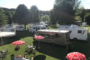 Wohnmobile und Campingbusse auf einer grünen Wiese, Tische und Stühle am Stellplatz, umgeben von hohen Bäumen.