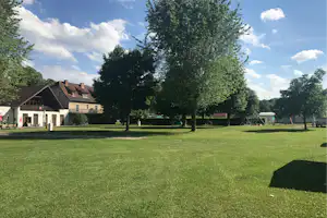 Grüne Campingwiese mit Bäumen, daneben Gebäude mit Terrasse und Sitzgelegenheiten, im Hintergrund Spielgeräte und Zäune.