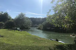 Blick auf den Campingsee mit grünem Ufer, Ruderbooten und Bojen im Wasser, umgeben von Bäumen und Wiese.