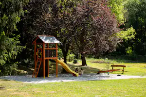 Spielplatz mit Kletterturm und gelber Rutsche auf dem Campingplatz, umgeben von Bäumen und Rasen.