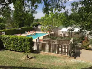 Schwimmbecken am Campingplatz mit Liegen, Hecken und Holzzaun; dahinter stehen weiße Wohnwagen und Bäume im Garten.