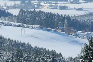 Winterlicher Campingplatz im Schnee mit Wohnwagen und Zelten am Hang, umgeben von verschneiten Bäumen und Häusern im Tal.