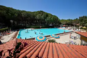 Camping-Pool mit türkisfarbenem Wasser, Sonnenliegen und Sonnenschirmen auf der Terrasse, im Hintergrund Bäume und Gebäude.