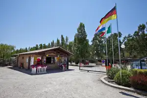 Campingplatz-Gebäude mit überdachter Terrasse, Sitzbereich und mehreren Flaggen vor dem Eingang, daneben Kiesweg und Hecken.