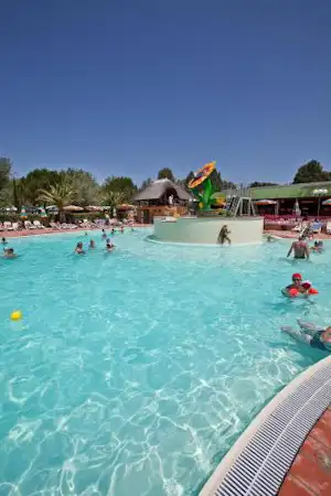 Kinder im Schwimmbecken vor einem großen Wasserspiel mit Fontäne, im Hintergrund Bäume und Campinganlage mit Liegen.