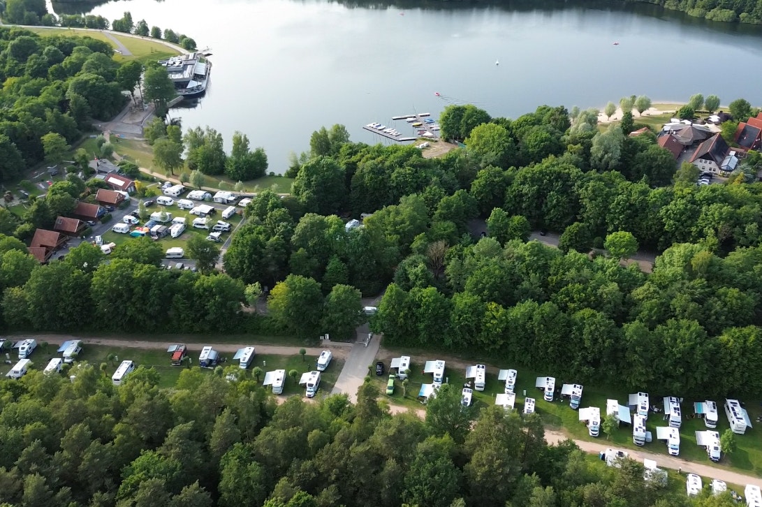 Campingplatz am See mit vielen Stellplätzen, Wohnmobilen und Bäumen, Blick von oben auf Ufer und Stege.