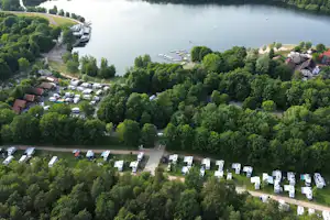 Campingplatz am See mit vielen Stellplätzen, Wohnmobilen und Bäumen, Blick von oben auf Ufer und Stege.