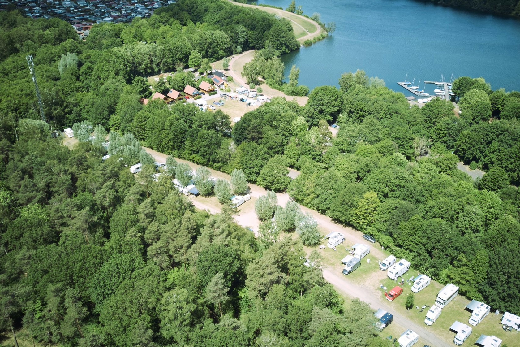 Luftaufnahme eines Campingplatzes am Fluss mit Stellplätzen, Wegen und Booten im Wasser, umgeben von viel Grün.