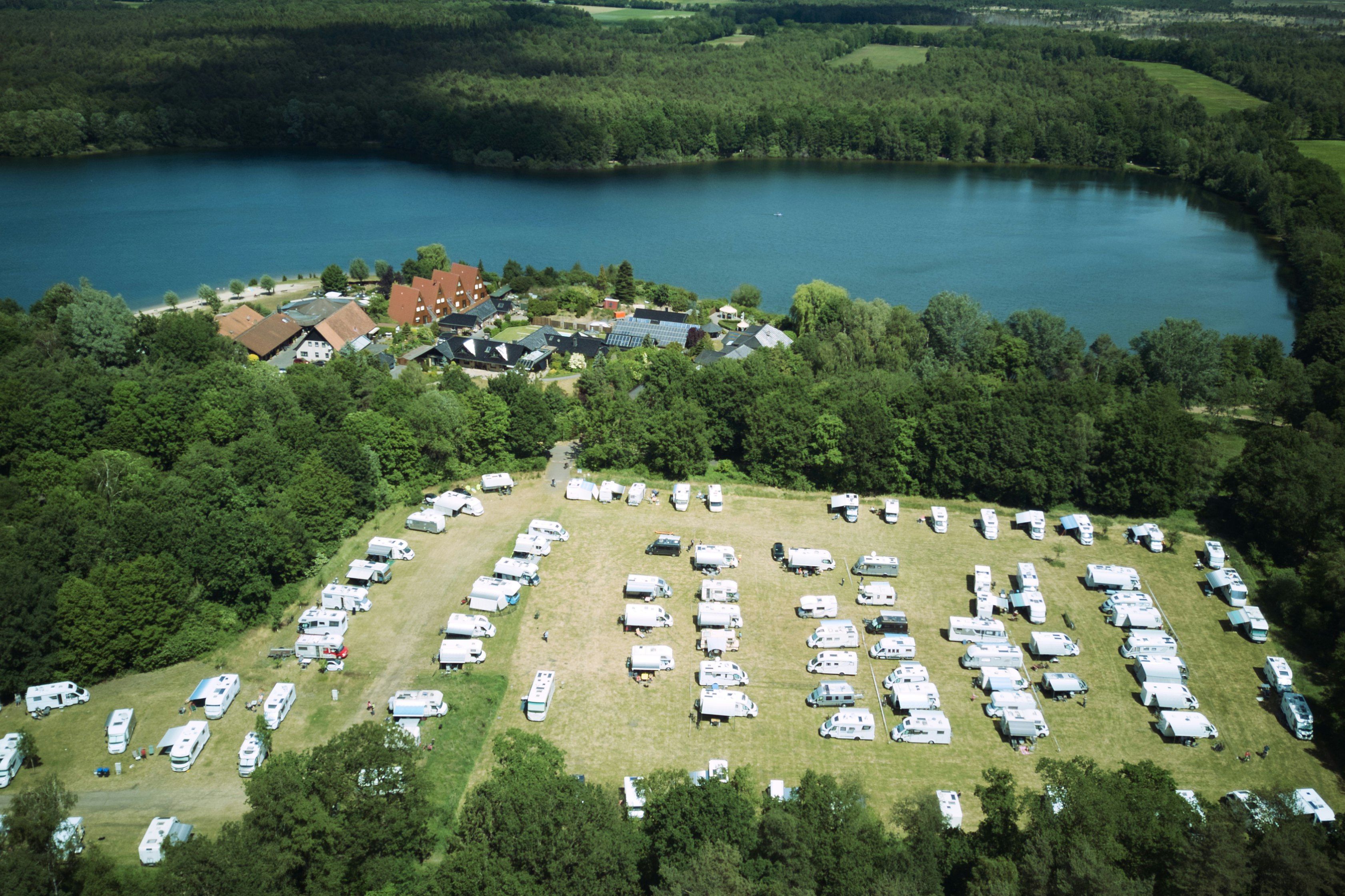 Campingplatz mit vielen Wohnwagen und Zelten auf einer Wiese, Blick auf den See und umliegende Bäume sowie Häuser im Hintergrund.