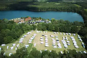 Campingplatz mit vielen Wohnwagen und Zelten auf einer Wiese, Blick auf den See und umliegende Bäume sowie Häuser im Hintergrund.