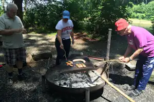 Männer grillen Würstchen und Gemüse an einem Feuerkorb auf einem Campingplatz, im Hintergrund Bäume und Kiesweg.