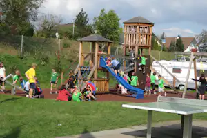 Holz-Spielplatz mit Rutsche und Kletterturm auf dem Campingplatz, daneben Wohnwagen und Autos, Rasen und Gehweg.