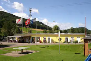 Campingplatz mit Wohngebäude, Rasenfläche und Sitzbereich, davor mehrere Flaggenmasten und Berge im Hintergrund.