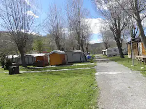 Campingplatz mit mehreren Zelten und einem Wohnwagen, Kiesweg zwischen Stellplätzen, Bäume und Rasen im Hintergrund.