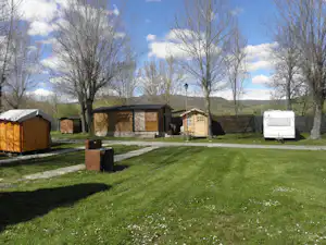 Grüner Rasen mit Wegen, daneben mehrere Gebäude und ein Wohnwagen auf einem Campingplatz, im Hintergrund Bäume und Wiese.