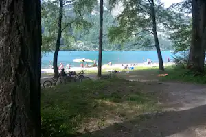 Campingplatz am See mit Liegewiese, Sonnenschirmen und vielen Menschen, im Vordergrund Bäume und ein Fahrrad.
