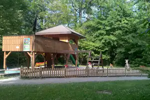 Spielhaus auf Stelzen mit Holzveranda, Schaukel und bunten Sitzbänken, umgeben von dichtem Wald und Zaun.