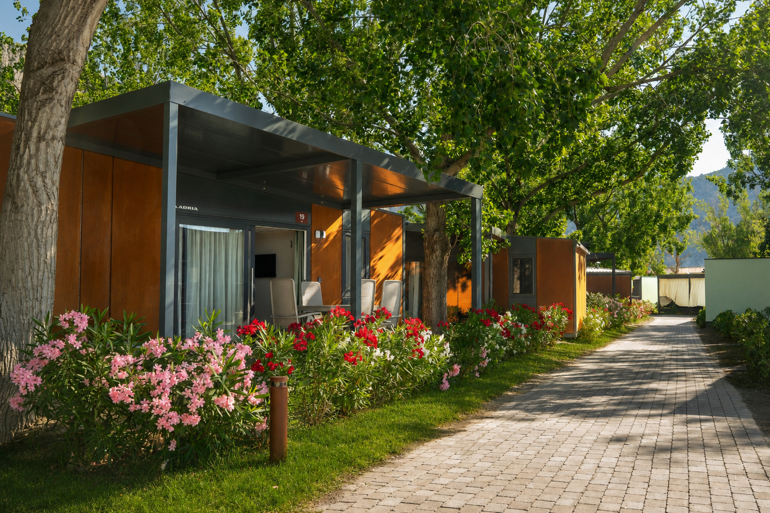 Reihen von Holzhäuschen mit Terrasse und Markise, gepflasterter Weg, Blumenbeete und Bäume im Hintergrund.