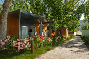 Reihen von Holzhäuschen mit Terrasse und Markise, gepflasterter Weg, Blumenbeete und Bäume im Hintergrund.