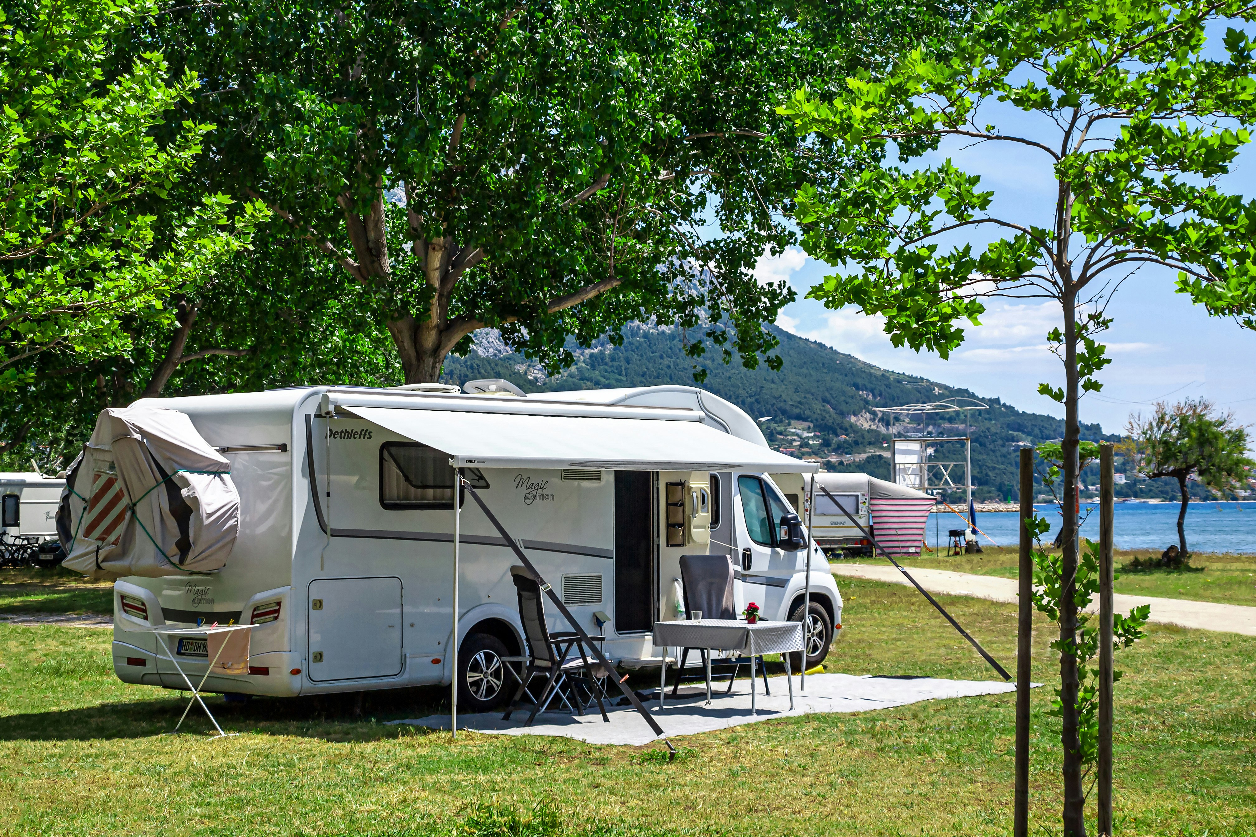 Wohnmobil mit Vorzelt und Campingstühlen auf einem Rasenplatz, umgeben von Bäumen und Blick auf das Wasser.