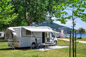 Wohnmobil mit Vorzelt und Campingstühlen auf einem Rasenplatz, umgeben von Bäumen und Blick auf das Wasser.