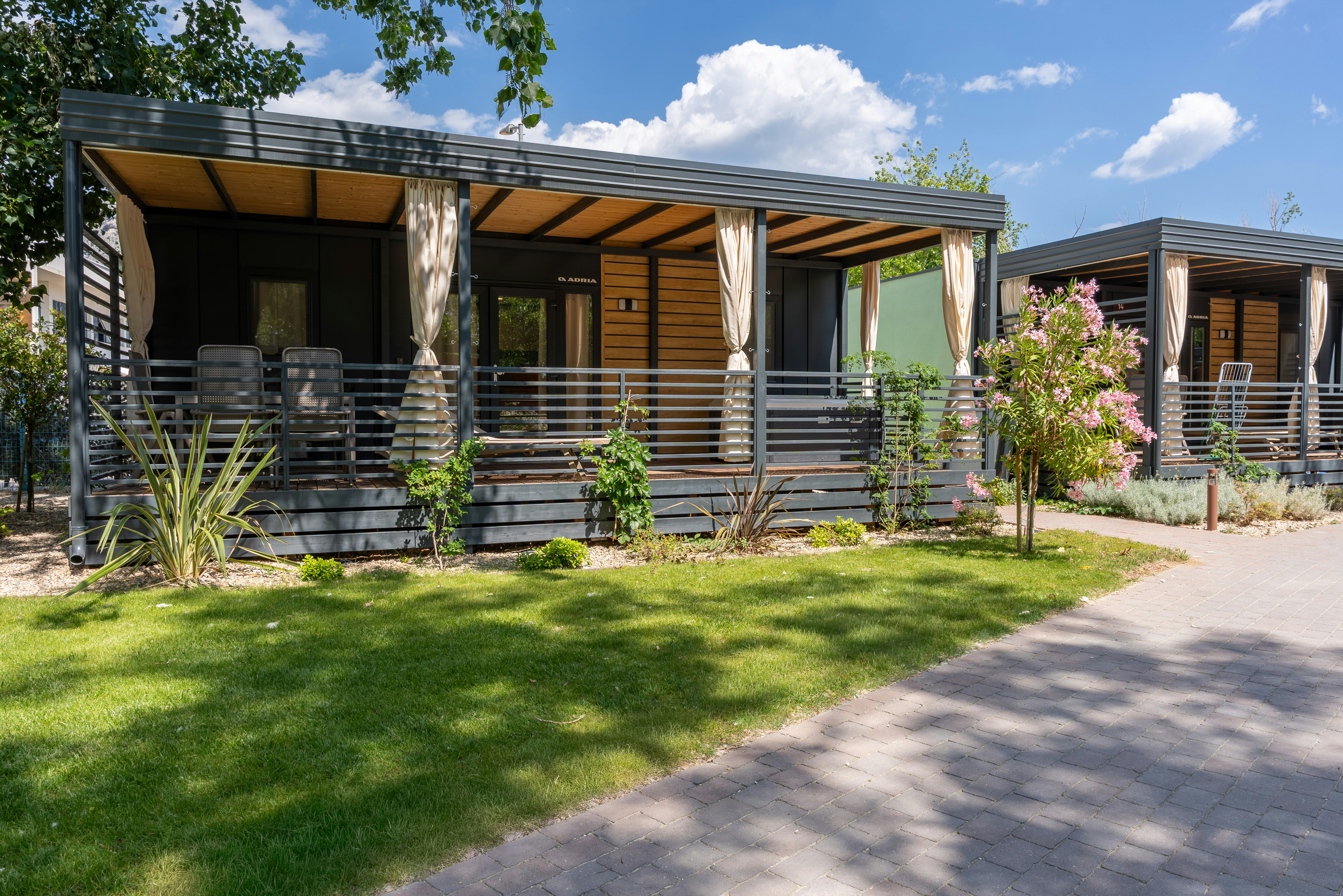 Holzbungalows mit überdachter Terrasse, weißen Vorhängen und Gartenrasen, daneben ein Kiesweg und Sträucher vor dem Gebäude.
