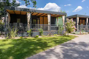Holzbungalows mit überdachter Terrasse, weißen Vorhängen und Gartenrasen, daneben ein Kiesweg und Sträucher vor dem Gebäude.