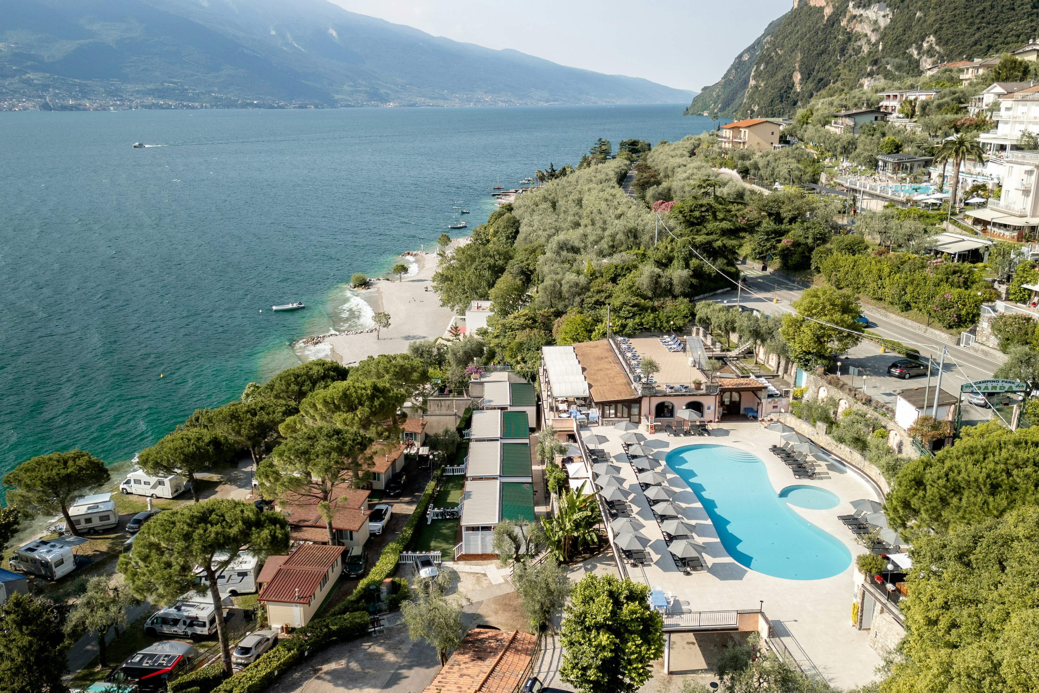 Campingplatz mit Pool am Hang, Blick auf den See und die Küste, Wohngebäude, Wege und geparkte Autos in der Nähe.