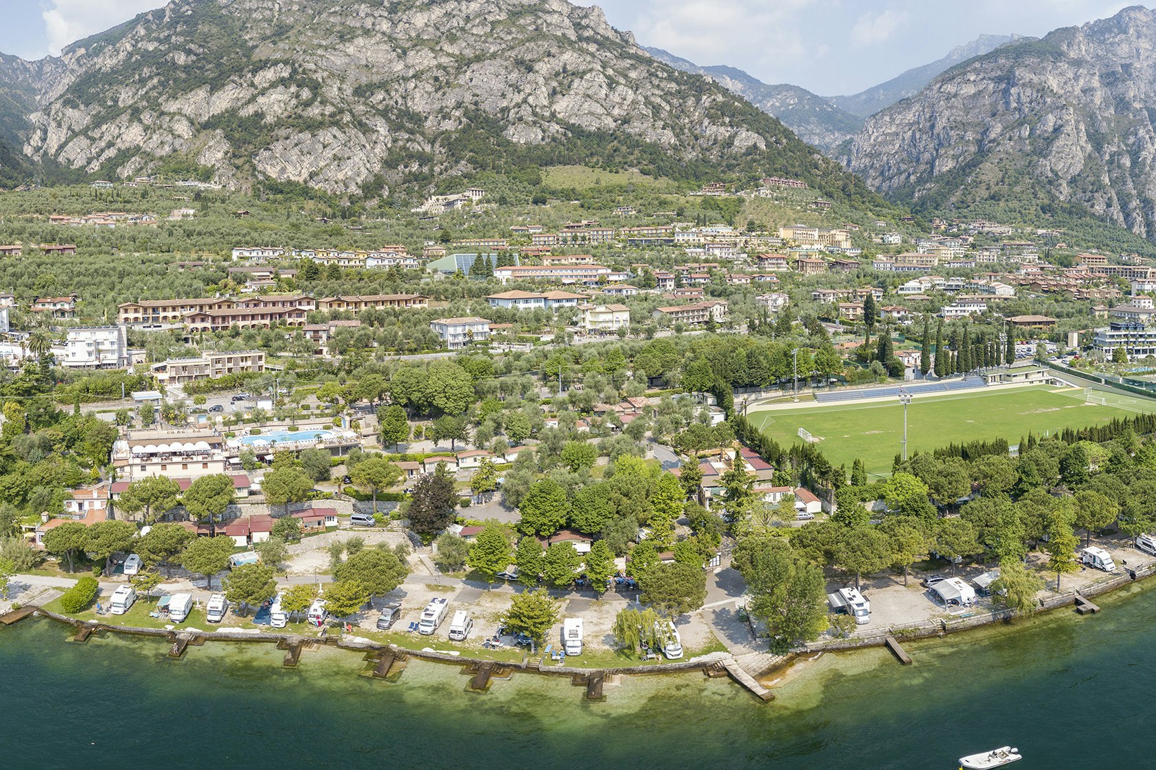 Luftaufnahme eines Campingplatzes am See mit Stellplätzen, Wohnwagen und Bäumen, umgeben von Bergen und einem Uferweg.