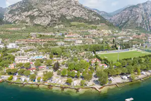 Luftaufnahme eines Campingplatzes am See mit Stellplätzen, Wohnwagen und Bäumen, umgeben von Bergen und einem Uferweg.