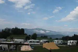 Campingplatz mit Wohnmobilen und Zelten, Blick auf grüne Wiesen und Berge in der Ferne unter wolkigem Himmel.