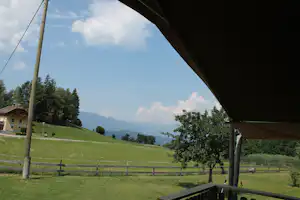 Blick von einer Campingwiese unter dem Zeltdach auf Berge, Wiesen und Bäume, im Hintergrund ein Haus am Hang.