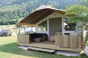 Campingzelt mit großer Markise und Holzterrasse, Sitzbank und Tisch, im Hintergrund Wiesen und bewaldete Hügel.