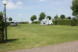 Campingwiese mit Wohnmobilen und Zelten, Hecken und Bäumen, gepflasterter Weg am Rand.