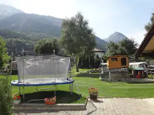 Trampolin im Campinggarten mit Rasen, Spielhaus und Sicht auf Berge und Bäume im Hintergrund.