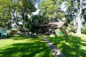 Campinghaus mit Terrasse, Gartenweg und Sitzgruppe vor dem Gebäude, umgeben von hohen Bäumen und Rasen.