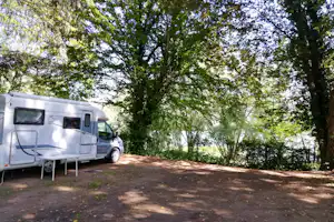 Wohnmobil auf einem Campingplatz am Waldrand, mit Vorzeltstangen und Stellplatz auf Schotter unter Bäumen.