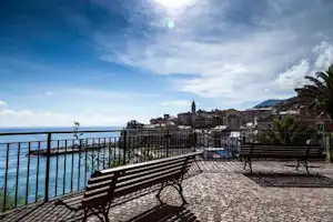 Terrasse mit Bank und Geländer, Blick aufs Meer und auf einen Küstenort mit Häusern und Palmen.