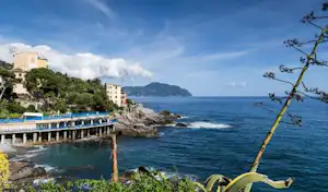 Campingplatz am Meer mit Steg und Küste, Felsen und Wasser, im Hintergrund Gebäude und bewachsene Hänge.