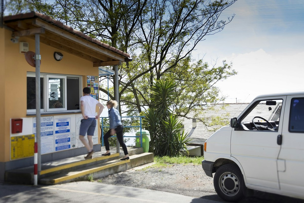 Campingplatz-Rezeption mit Informationswand, zwei Personen am Eingang und weißem Auto auf dem Stellplatz.