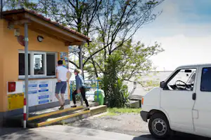 Campingplatz-Rezeption mit Informationswand, zwei Personen am Eingang und weißem Auto auf dem Stellplatz.