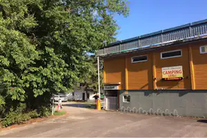Camping-Gebäude mit Eingangsschild, Holzfassade und Balkon, davor Asphaltweg und Bäume am Rand.