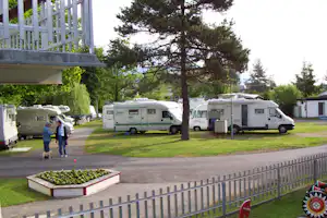 Wohnmobile auf dem Campingplatz, gepflasterter Weg und Rasenfläche mit Bäumen, im Hintergrund weitere Fahrzeuge und Gebäude.