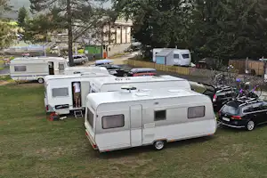 Mehrere Wohnwagen und ein Wohnmobil auf einem Campingplatz-Stellplatz, mit Rasen, Hecke, Fahrrädern und Bäumen im Hintergrund.